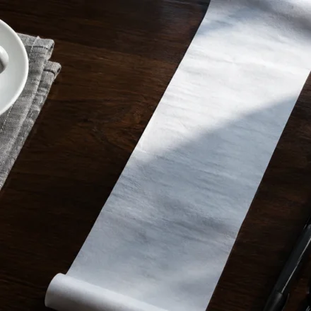 A long blank paper receipt on a dark walnut desk, half lit by a shaft of cool daylight and half in shadow, with an espresso cup, a pen and folded reading glasses beside it.