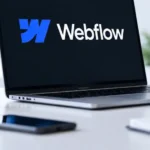 A laptop showing the Webflow logo on a bright desk beside a plant, a stack of books and a coffee mug